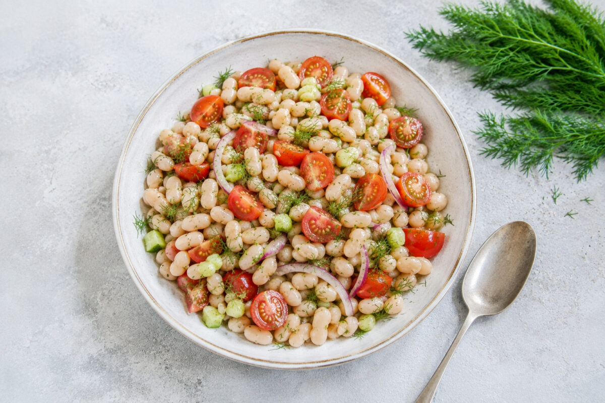 Fertiger Weiße-Bohnen-Salat aus deutlich höherer Perspektive in heller Keramikschale mit Tomaten, Gurke, roten Zwiebeln und Dill.