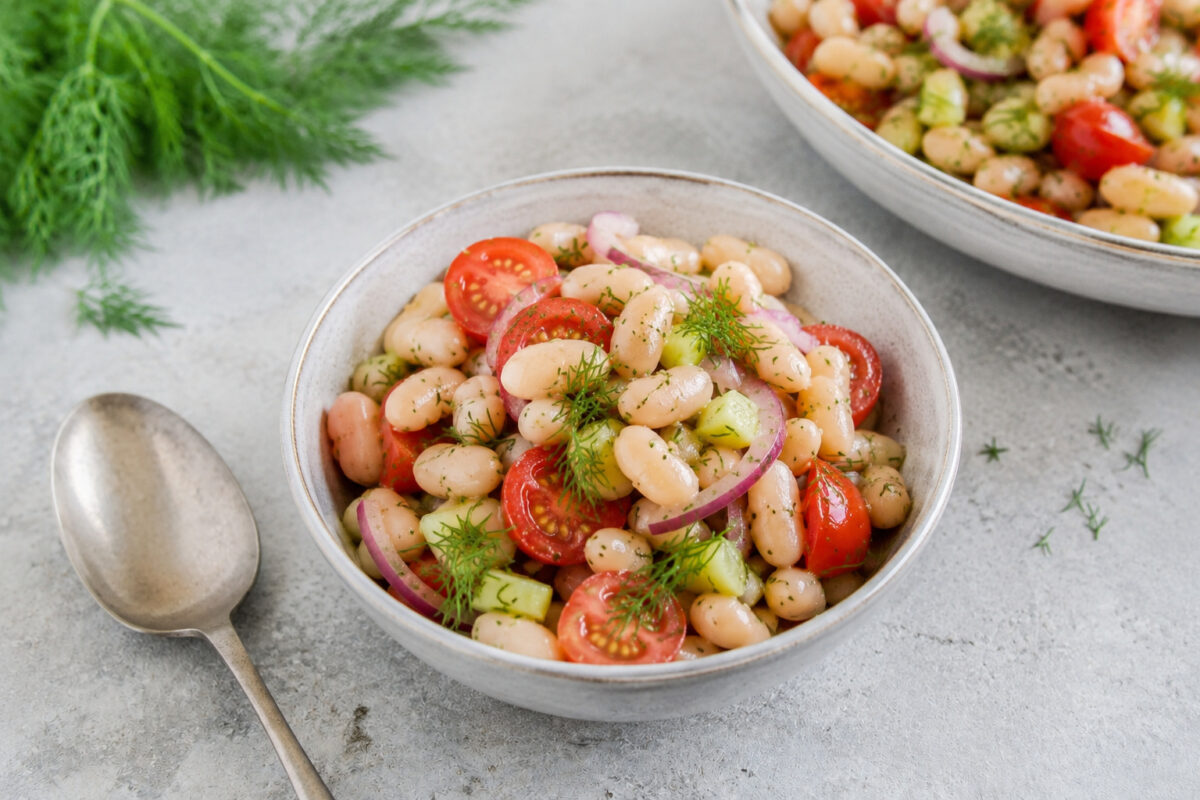 Servierszene mit einer kleinen Portionsschale des fertigen Weiße-Bohnen-Salats; größere Schale im Hintergrund, Dill und Löffel als Requisiten.