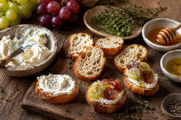 Zutaten- und Vorbereitungsansicht mit Baguette, Ziegenfrischkäse, roten und grünen Trauben, Honig, Olivenöl und Thymian.