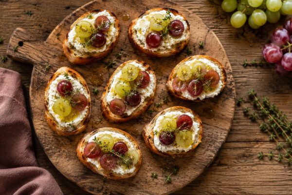 Draufsicht auf fertige Ziegenkäse-Crostini mit roten und grünen Trauben, Honig und Thymian.