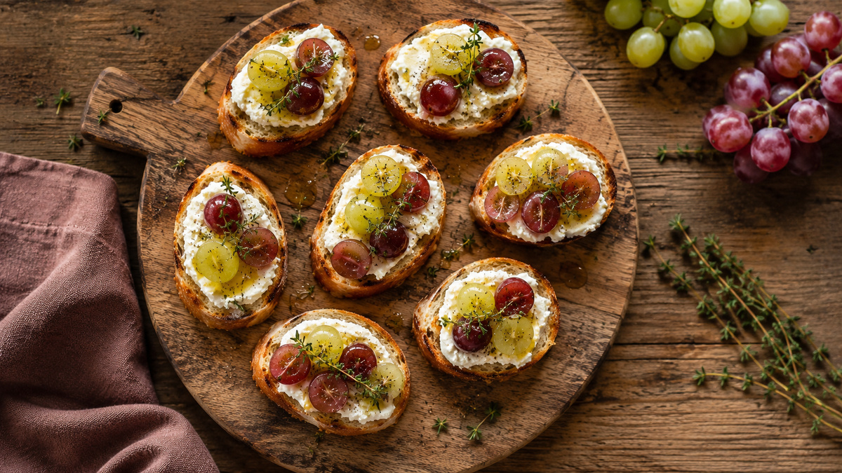 Draufsicht auf fertige Ziegenkäse-Crostini mit roten und grünen Trauben, Honig und Thymian.